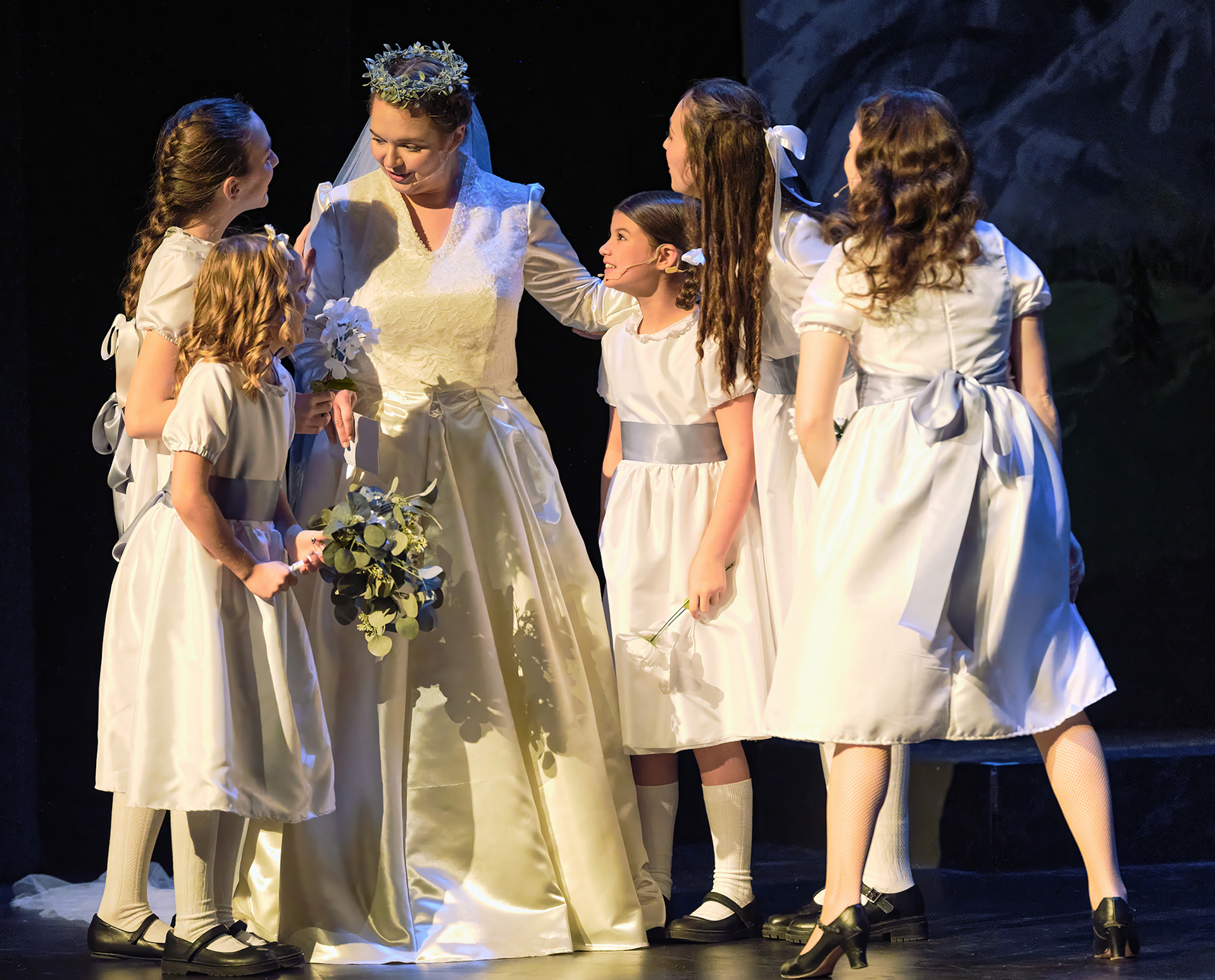 A woman in a wedding dress is surrounded by flower girls on a stage.