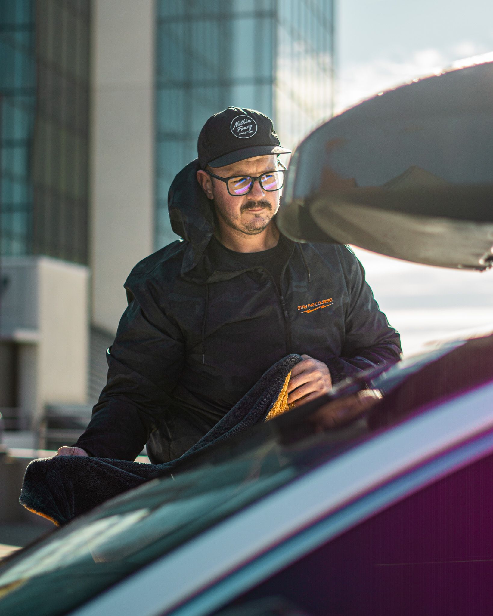 Man in black jacket and cap leans on a car, city background.