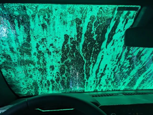 Inside a car at a carwash, green soap covers the windshield, obscuring the view.