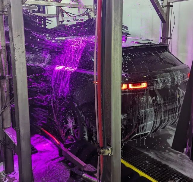 Car being washed in a car wash, black vehicle surrounded by purple-lit water and brushes.