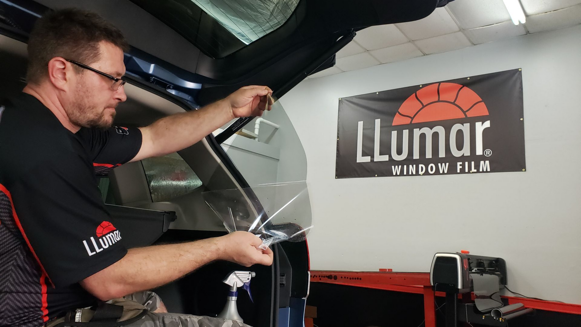 Man applying window tint to a car in a shop, LLumar sign in background.