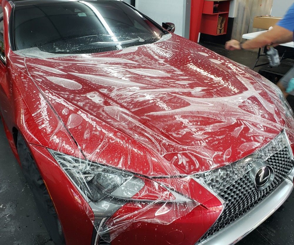 Red Lexus sedan covered in clear protective film in a workshop, with a person working nearby