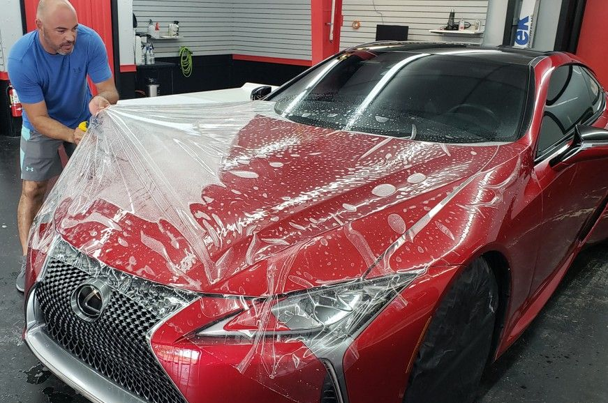 Man applying clear protective film to a red Lexus car hood in a garage.