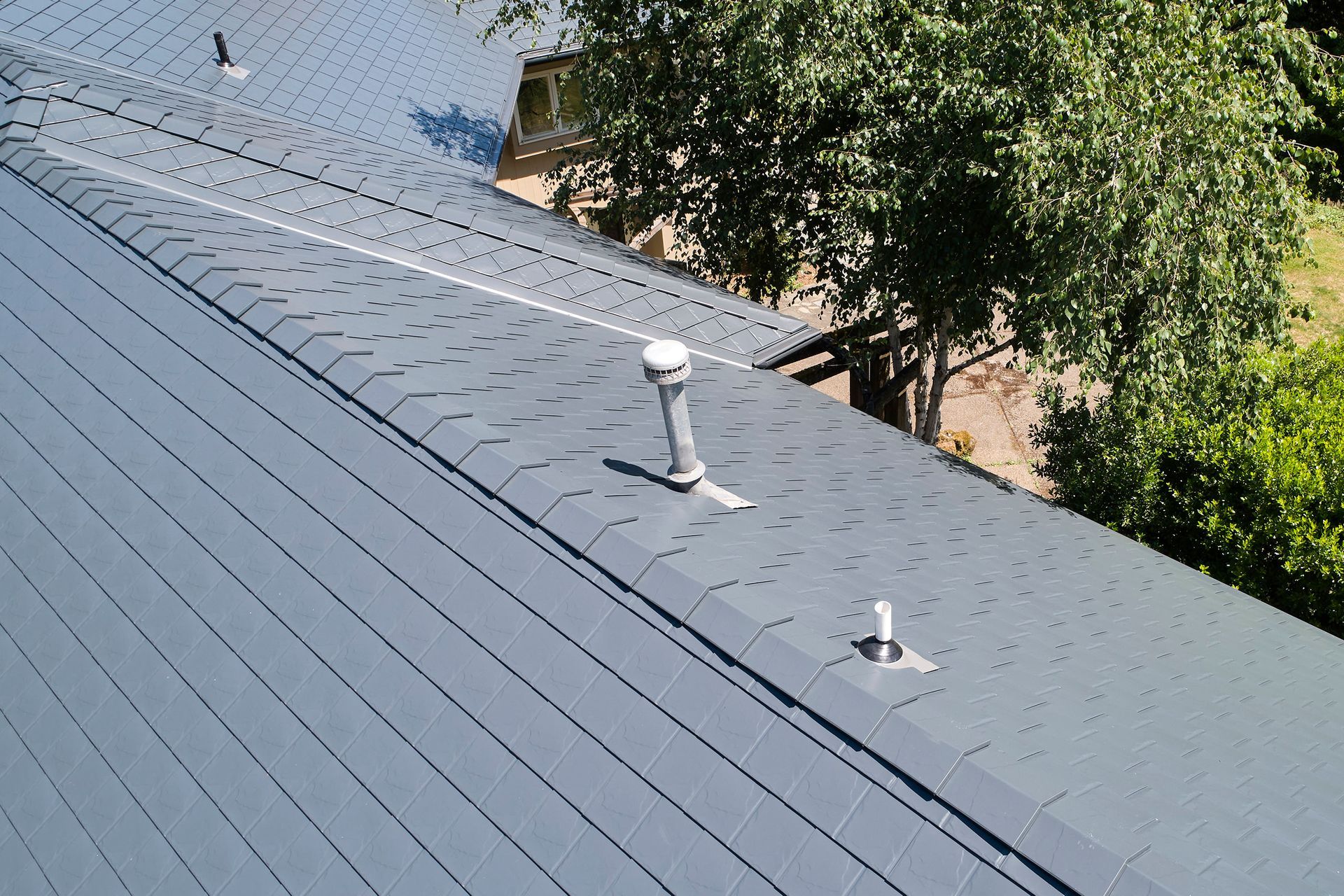 Aerial shot of an estate roofline with prominent brick chimneys and a Deep Charcoal Interlock Slate aluminum roof in Portland’s Dunthorpe neighborhood. The fire-resistant aluminum roof contrasts beautifully against the wooded backdrop, offering low-maintenance protection with upscale curb appeal.
