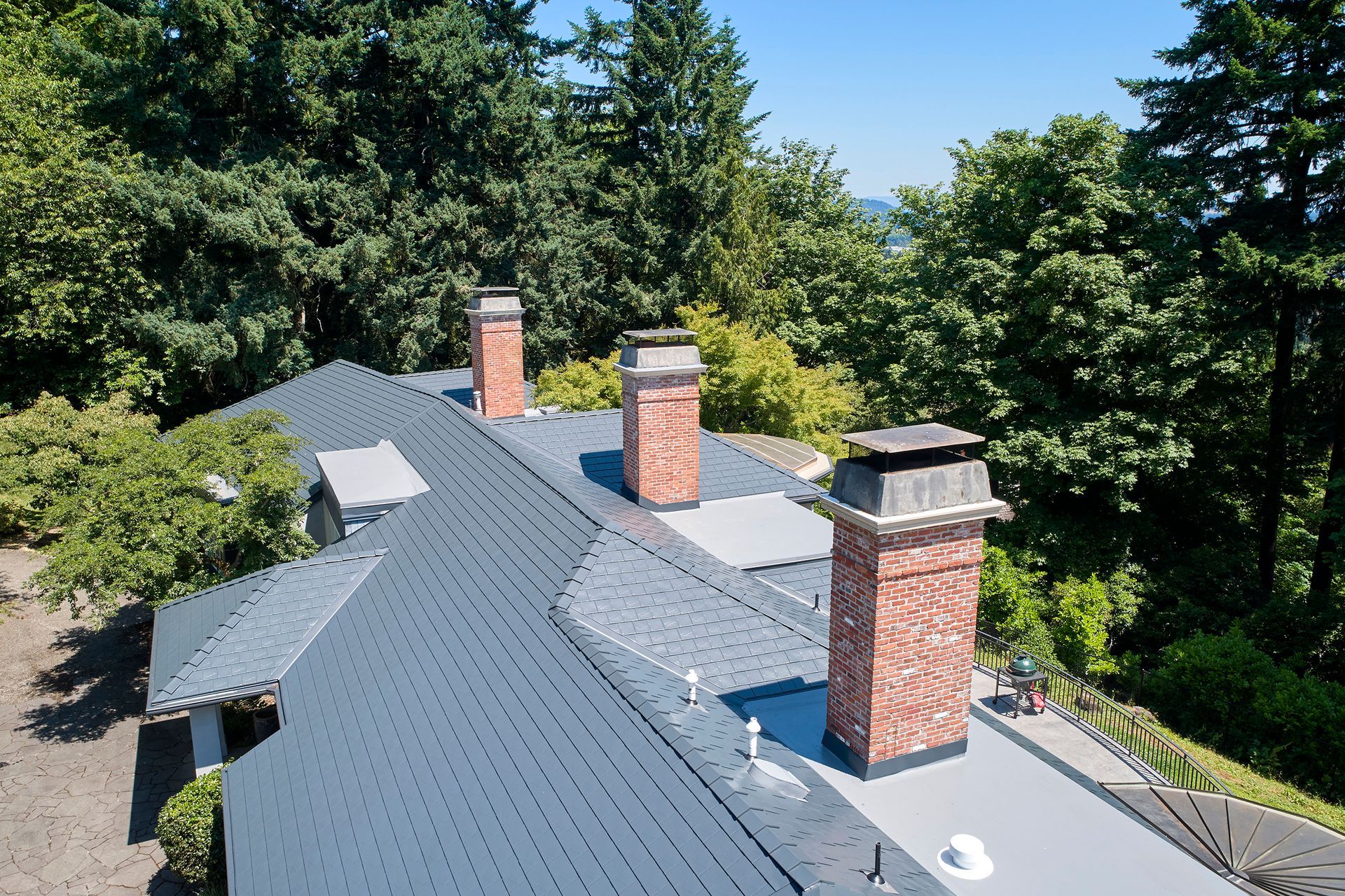 Architectural shot of multiple red brick chimneys emerging from a modern deep charcoal Interlock aluminum slate roof in the Dunthorpe neighborhood of Portland, Oregon. The metal slate shingles are installed with precision, ensuring long-term performance and visual impact.