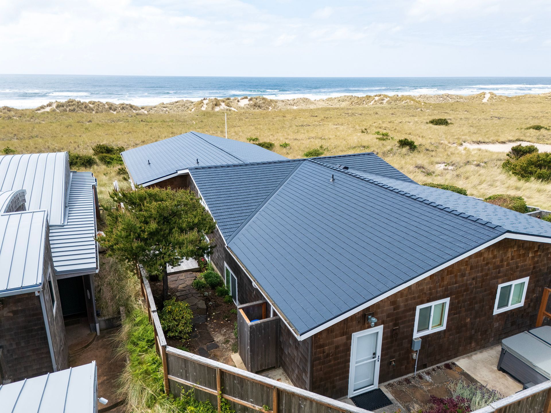 Aerial view of a beachfront home in the Buena Vista neighborhood of Manzanita, Oregon, featuring a Black Interlock Cedar Shingle Roof installed by Interlock Metal Roofing. The heavy-gauge aluminum roofing system is engineered with a 4-way interlocking design, providing unmatched resistance to high coastal winds typical of Manzanita Beach. The custom-fitted black shingles offer superior protection against salt spray thanks to their advanced Alunar® Coating, while the home’s architecture includes cedar shake siding, white trim, a coastal deck, and panoramic views of sand dunes and the Pacific Ocean.