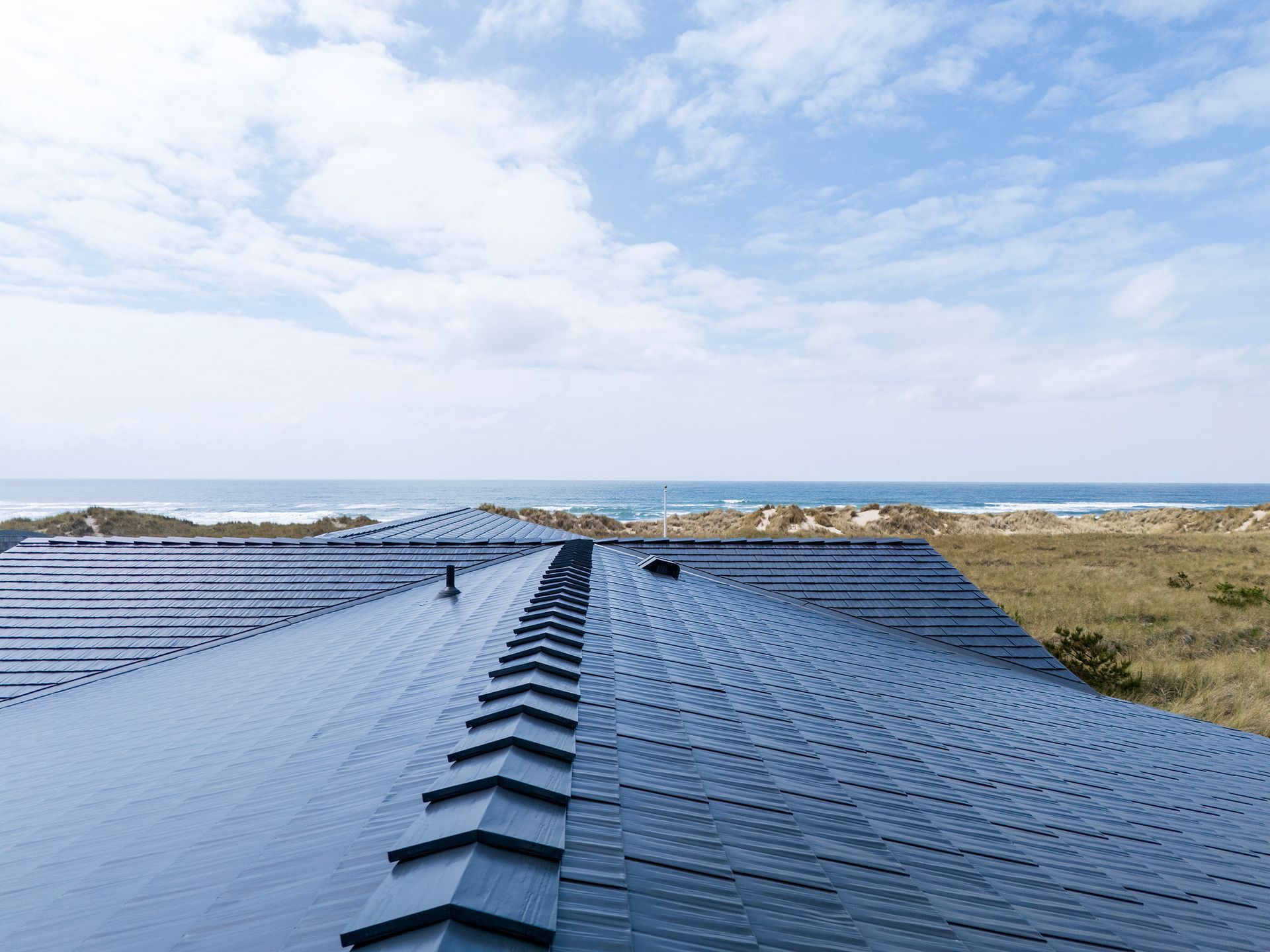 Close-up ridge view of a Black Interlock Cedar Shingle Roof installed on a coastal home in Manzanita, Oregon, highlighting the clean geometric precision of the patented 4-way interlocking aluminum shingle system. This elevated angle looks west toward the Pacific Ocean, showing how the weather-resistant aluminum roofing, coated with Alunar®, withstands salt-laden air and high wind exposure common to the Tillamook County coastline. The seamless ridge cap and interlocking seams emphasize storm-grade durability and year-round protection for Oregon beach homes in extreme marine environments.