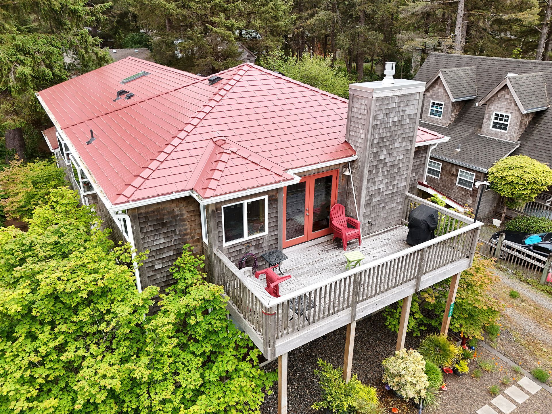 Aerial view of a coastal cedar-sided beach home in Manzanita, Oregon, highlighting a freshly installed Tile Red Interlock Cedar Shingle Roof by Interlock Metal Roofing. The metal shingles, made of durable heavy gauge aluminum, are anchored with a four-way interlocking system that offers exceptional wind resistance against Manzanita Beach’s notorious gusts. The Alunar® coating ensures long-term performance by protecting against rust and salt air corrosion. This contemporary beach house features a central chimney, red-trimmed double doors, skylights, and strategically placed vents that blend utility with aesthetics. The elevated wood deck provides a panoramic view and connects seamlessly with the roofline, showcasing Interlock’s craftsmanship in protecting homes in harsh coastal environments.
