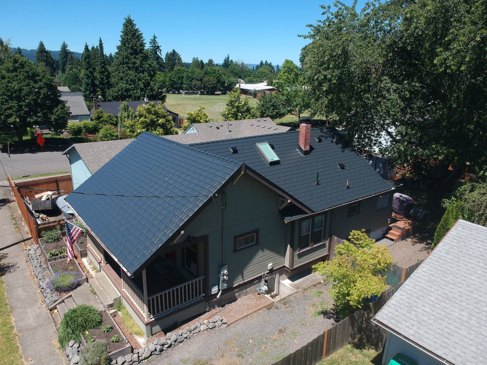 Historic Craftsman-style home in the St. Johns neighborhood of Portland, Oregon, featuring a premium Black Interlock Cedar Shingle Roof made from heavy-gauge, rust-resistant aluminum installed by Interlock Metal Roofing. The architectural character of the home includes deep overhanging eaves, exposed rafter tails, a gabled roof with a prominent front-facing porch, a red brick chimney, and a central skylight that enhances natural light indoors. The durable aluminum shingles are engineered with a 4-way interlocking design to provide superior wind uplift resistance, essential for homes in Portland’s changing weather patterns. The Alunar® coating system offers long-lasting protection from rain, moss, and UV degradation, preserving the deep black color for decades. The landscape includes a raised garden bed, flagstone retaining wall, mature tree canopy, and patriotic American flag—complementing the home’s Northwest charm and enhancing curb appeal in one of Portland’s most character-rich communities.