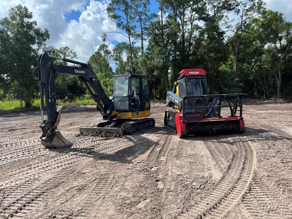 Excavator and brush cutter on a cleared dirt lot. Trees in background.
