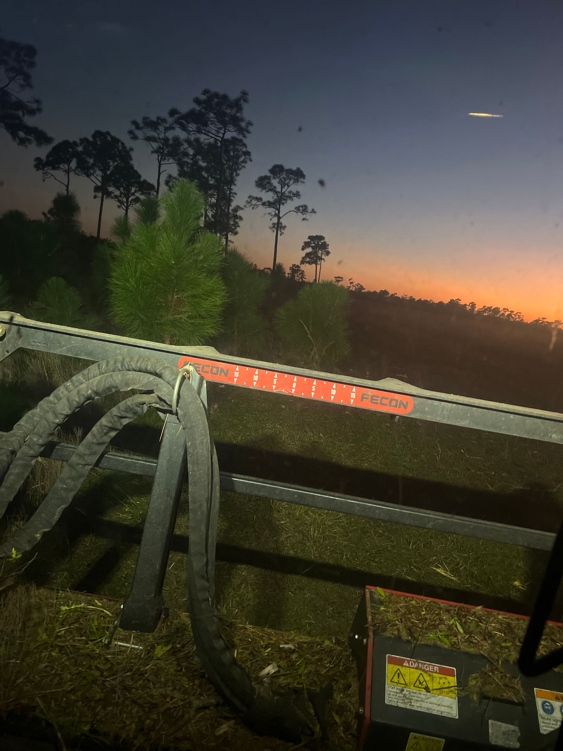 View from a tractor cab at sunset; trees line a distant hill.