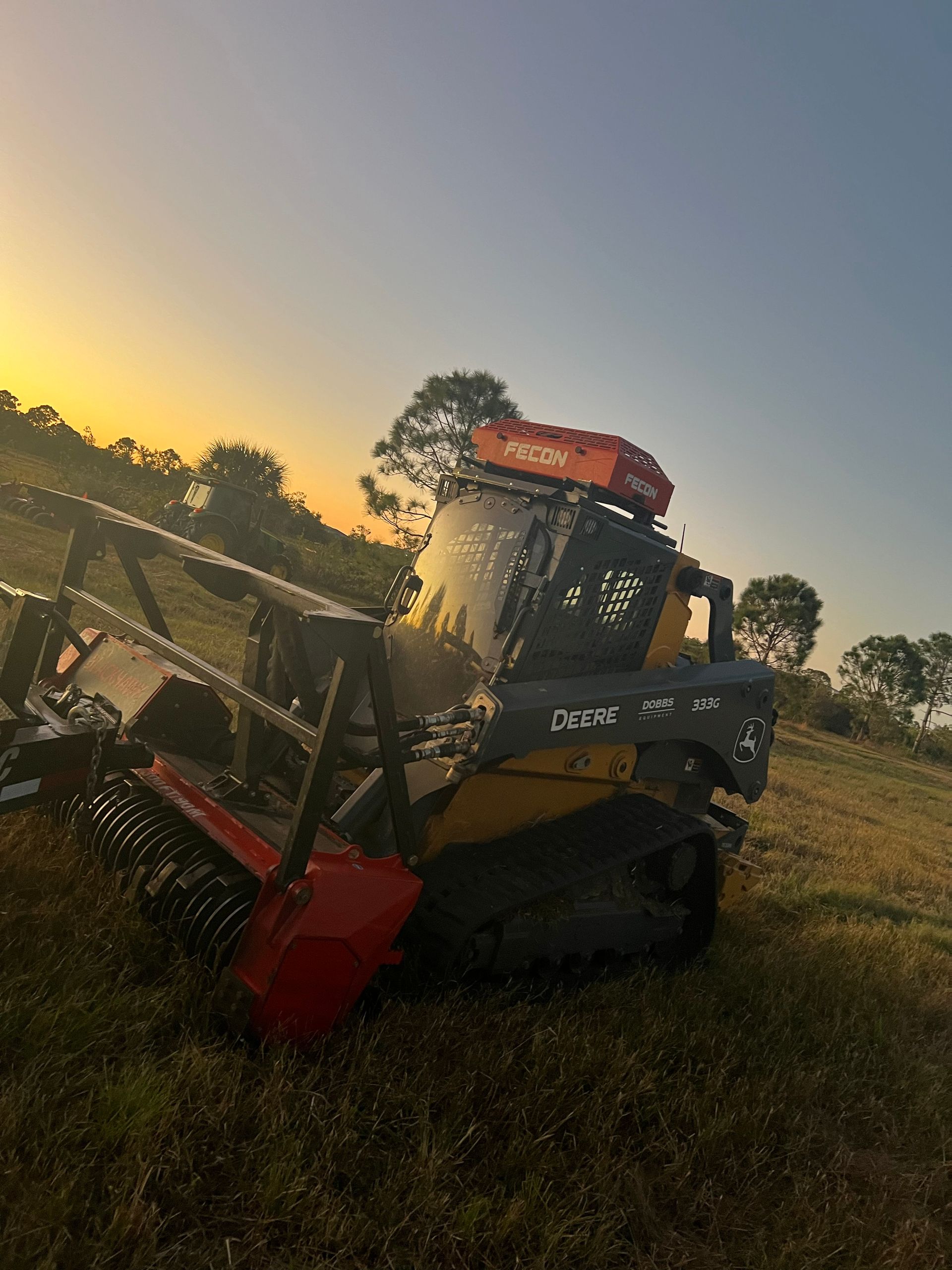 A small, yellow and black skid steer with a red brush attachment is on a grassy hill at sunset.