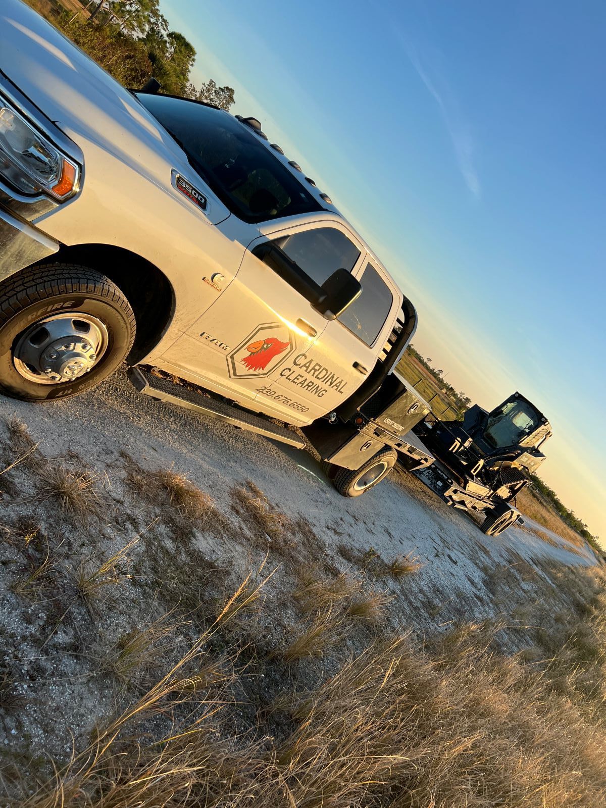 White work truck hauling a small excavator on a gravel road, under a clear sky.