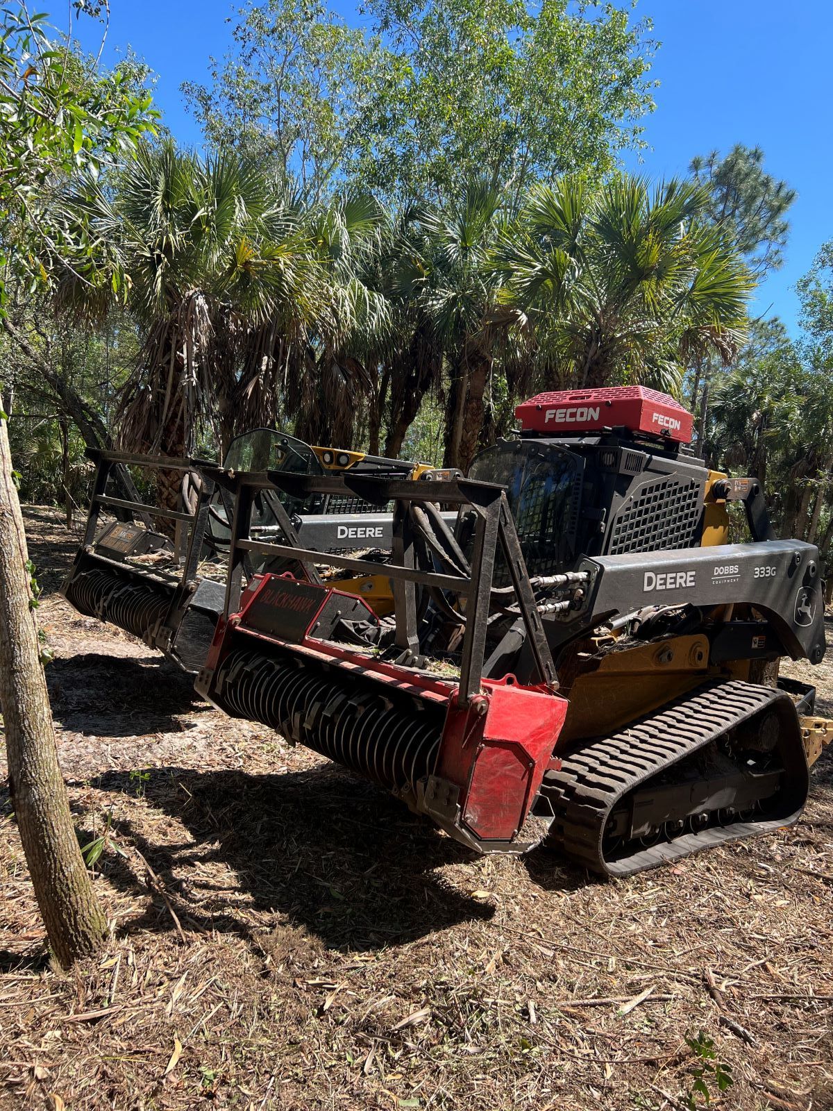 A tracked forestry mulcher in a wooded area, with red and black equipment, blue sky.