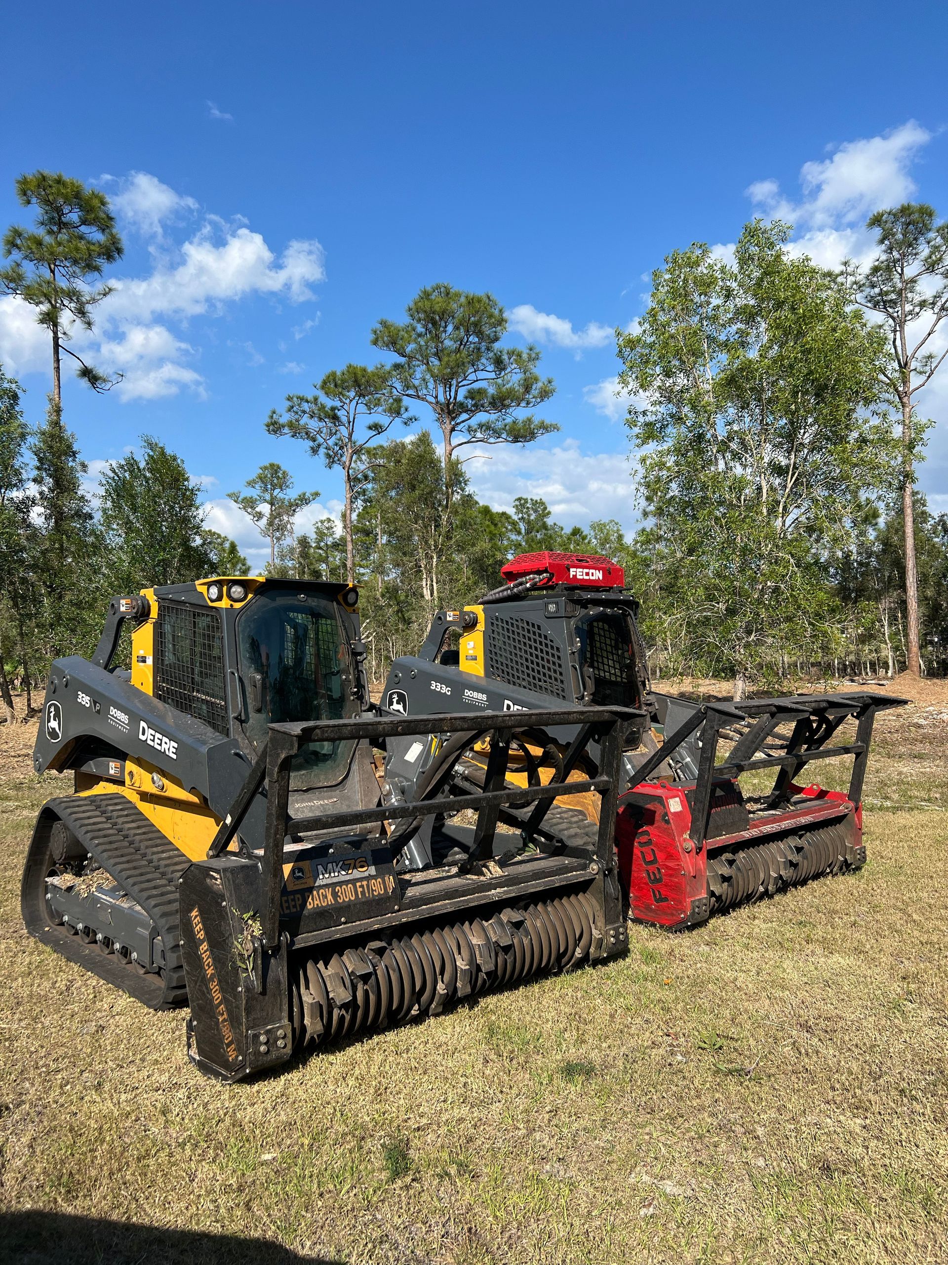 Two yellow and black forestry mulchers on tracks, in a sunny field with trees.