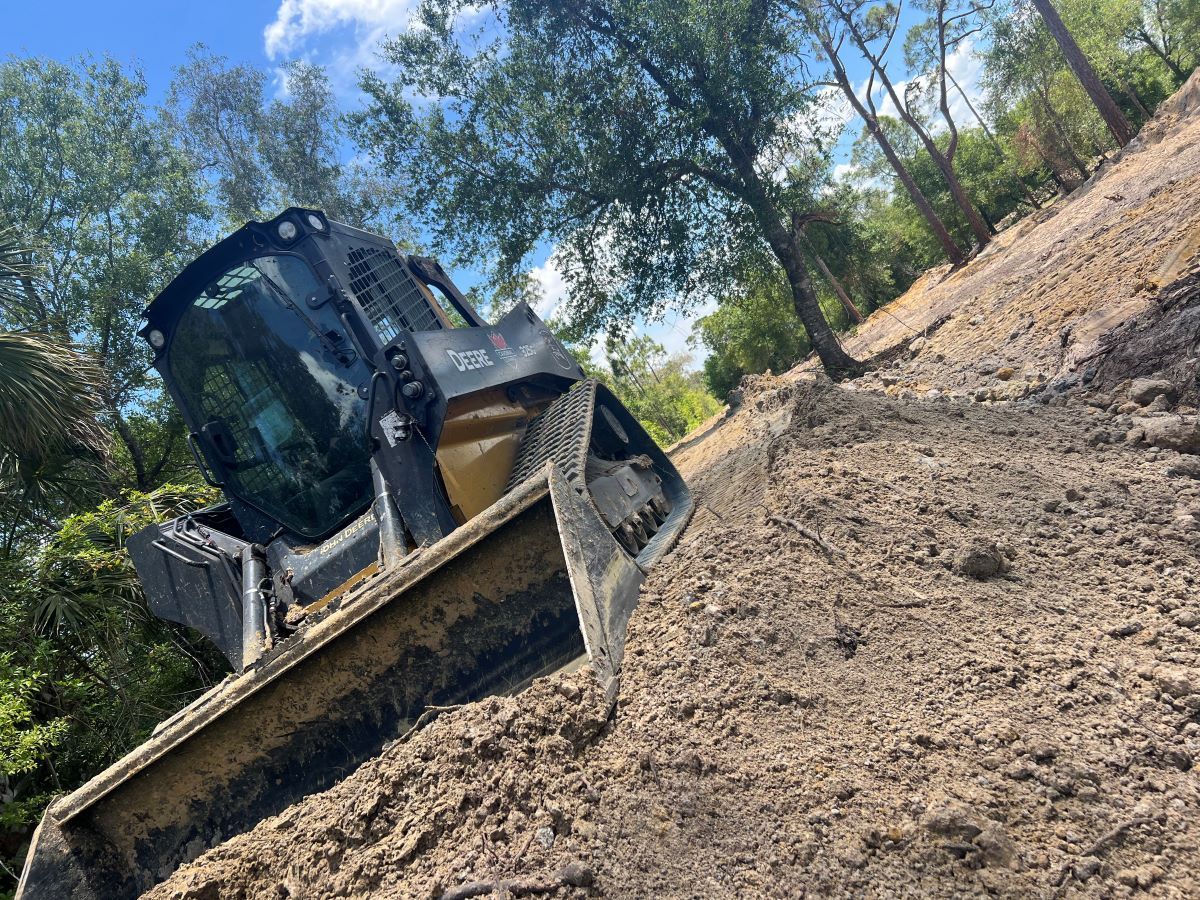 Skid steer on a dirt road, grading the terrain. Cloudy sky and trees in the background.