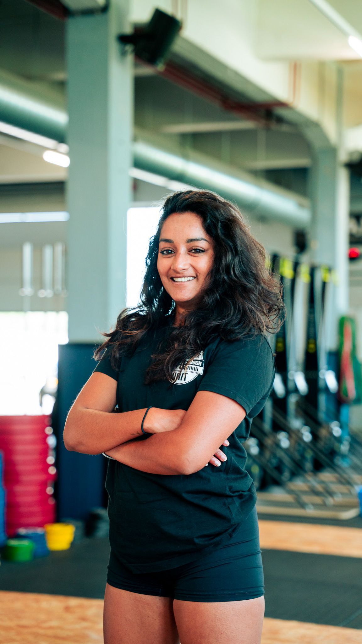 Een vrouw staat in een sportschool met haar armen gekruist en glimlachend.