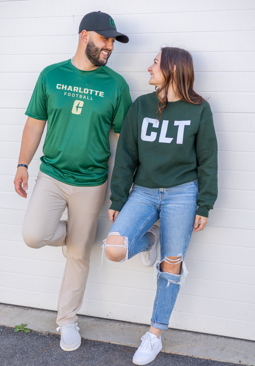 Couple wearing green Charlotte apparel, smiling and leaning against a white wall.