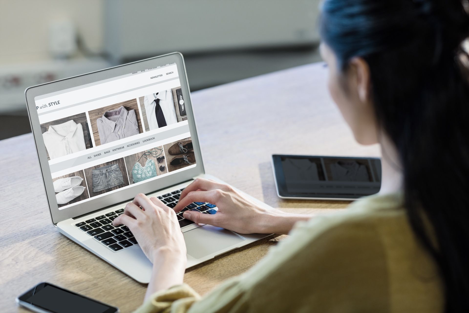 Person seated at table using a laptop, shopping online. Phone and tablet are also on the table.