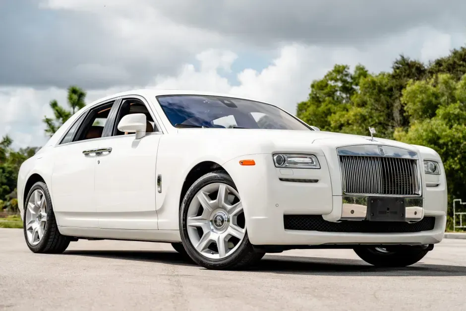 White Rolls-Royce Ghost luxury sedan parked on a paved road, under a bright sky and trees.