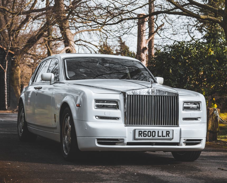 White Rolls-Royce Phantom luxury car parked on a road. Silver grille, clear sky, trees in background.