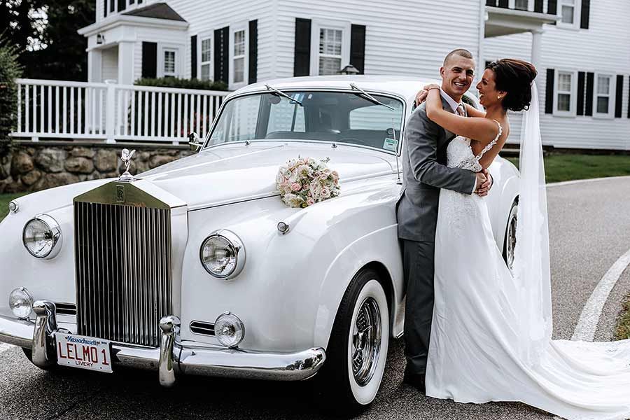 Newlyweds embrace next to a white vintage Rolls Royce. Bride in a white dress, groom in gray suit. Exterior shot.