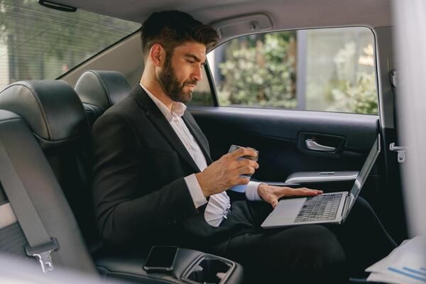 Businessman in a suit working on a laptop in the back seat of a car, holding a drink.