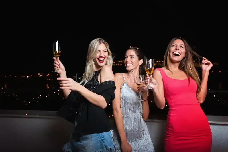 Three women laughing and toasting champagne glasses on a balcony at night, city lights in background.