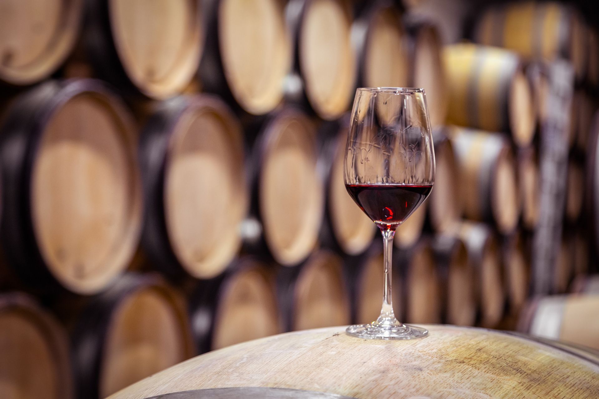 A glass of red wine is sitting on top of a wooden barrel in a wine cellar.