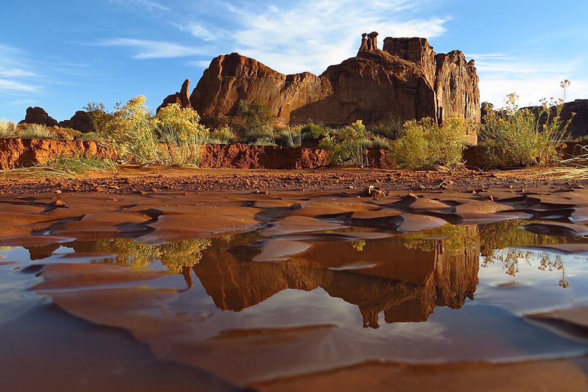 Red rock formation reflected in a muddy puddle, under a blue sky.