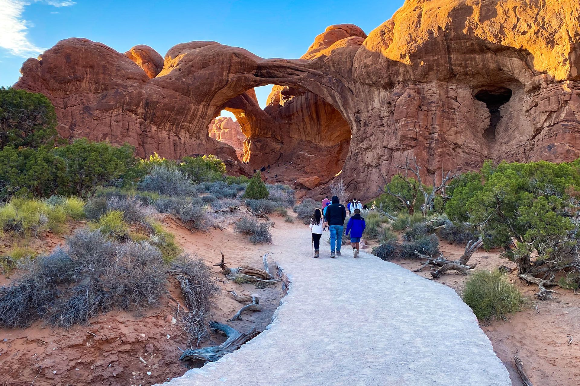 People walking on a path toward sandstone arches in a desert landscape.