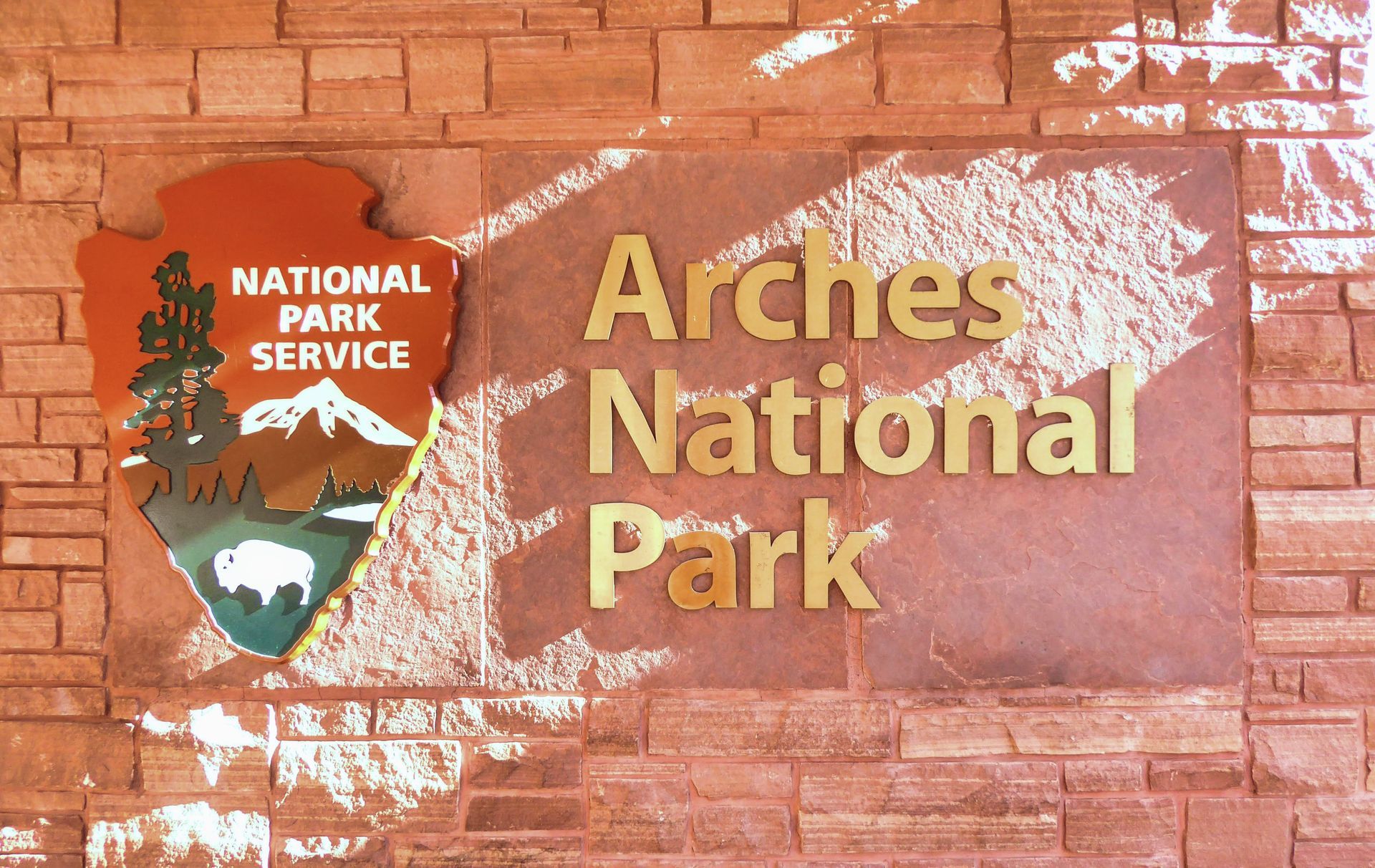 Sign for Arches National Park, with the National Park Service logo and gold lettering on a brick wall.