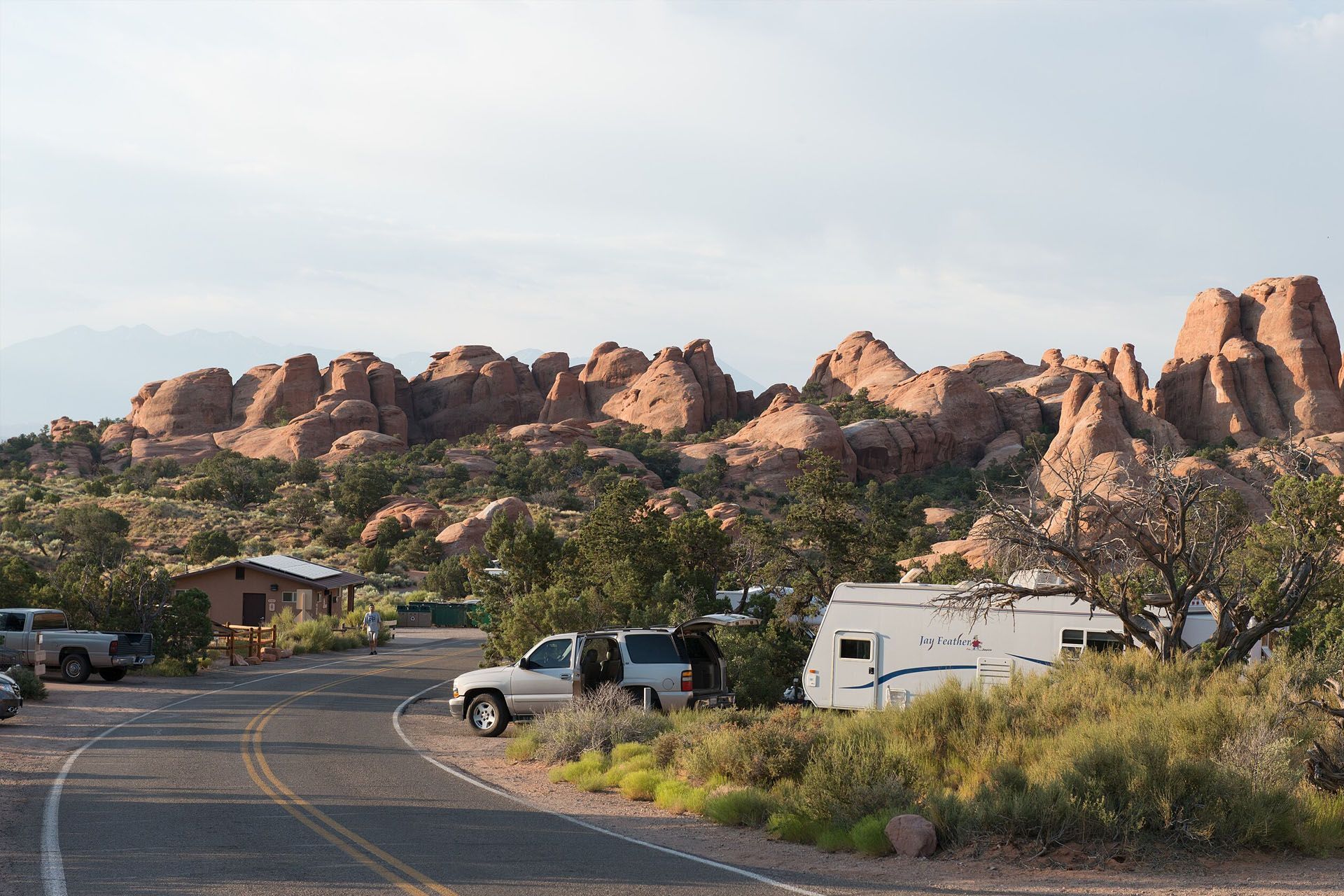 Campsite with RV and truck along a road, reddish rock formations in the background under a cloudy sky.