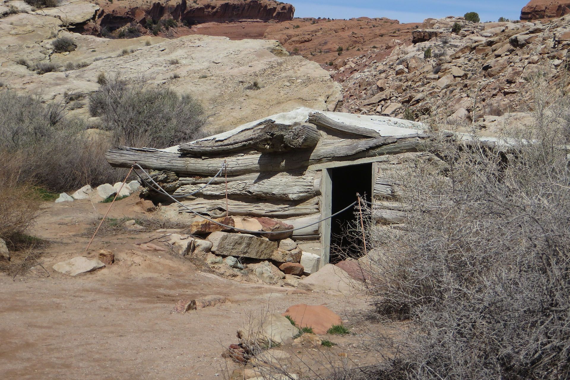 Small, weathered log cabin with open doorway in a desert landscape. Brown and tan colors.