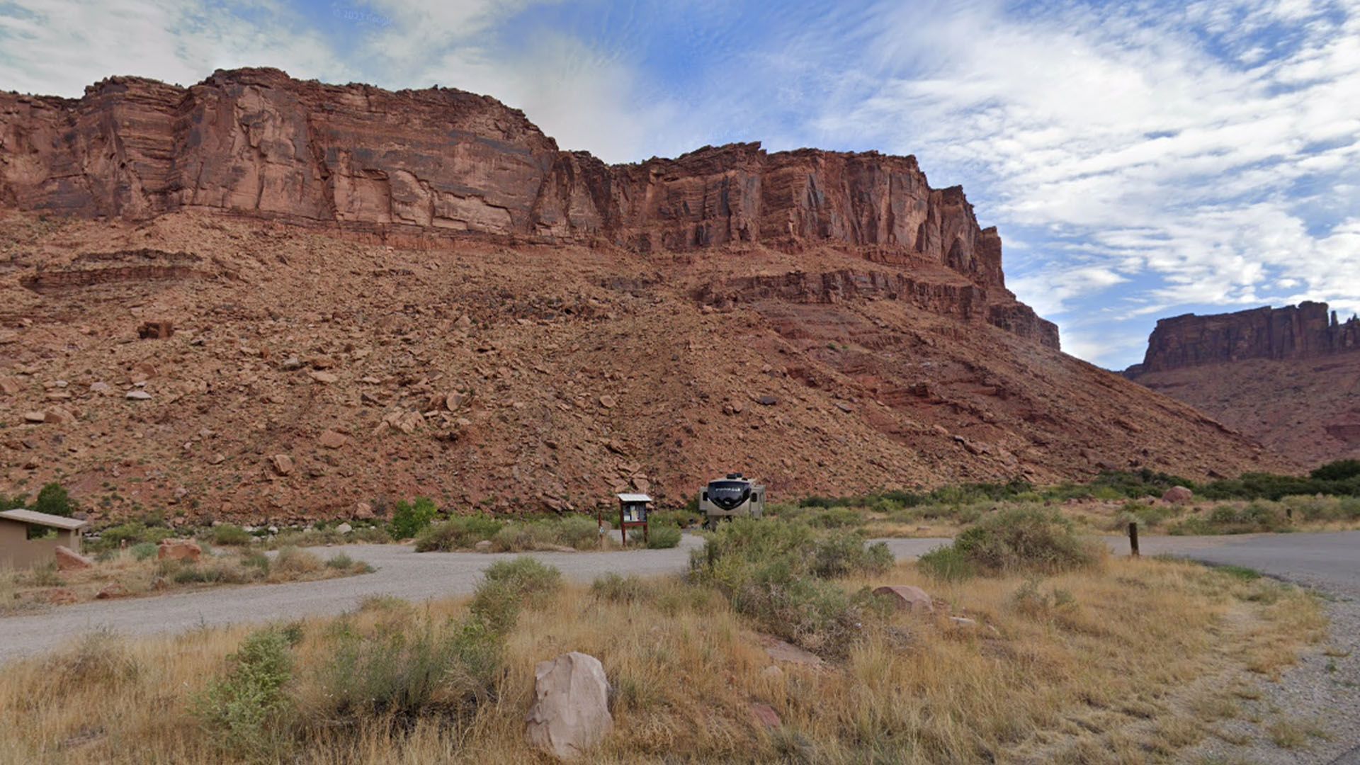 Campsite at base of large red rock cliffs; vehicles, dry vegetation, and a partly cloudy sky.