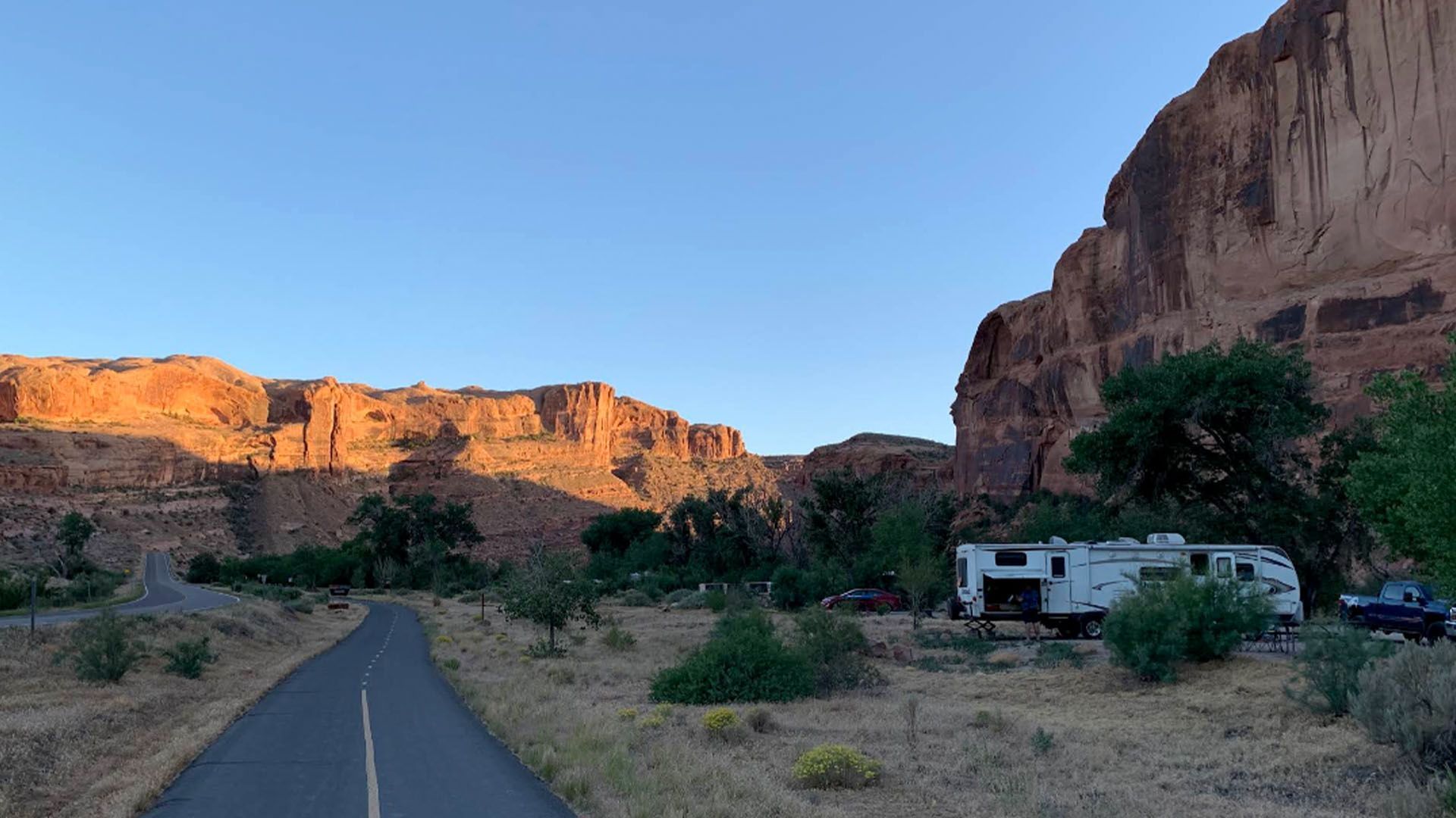 RV parked near a canyon, road in foreground, golden sunlight on canyon walls, blue sky.