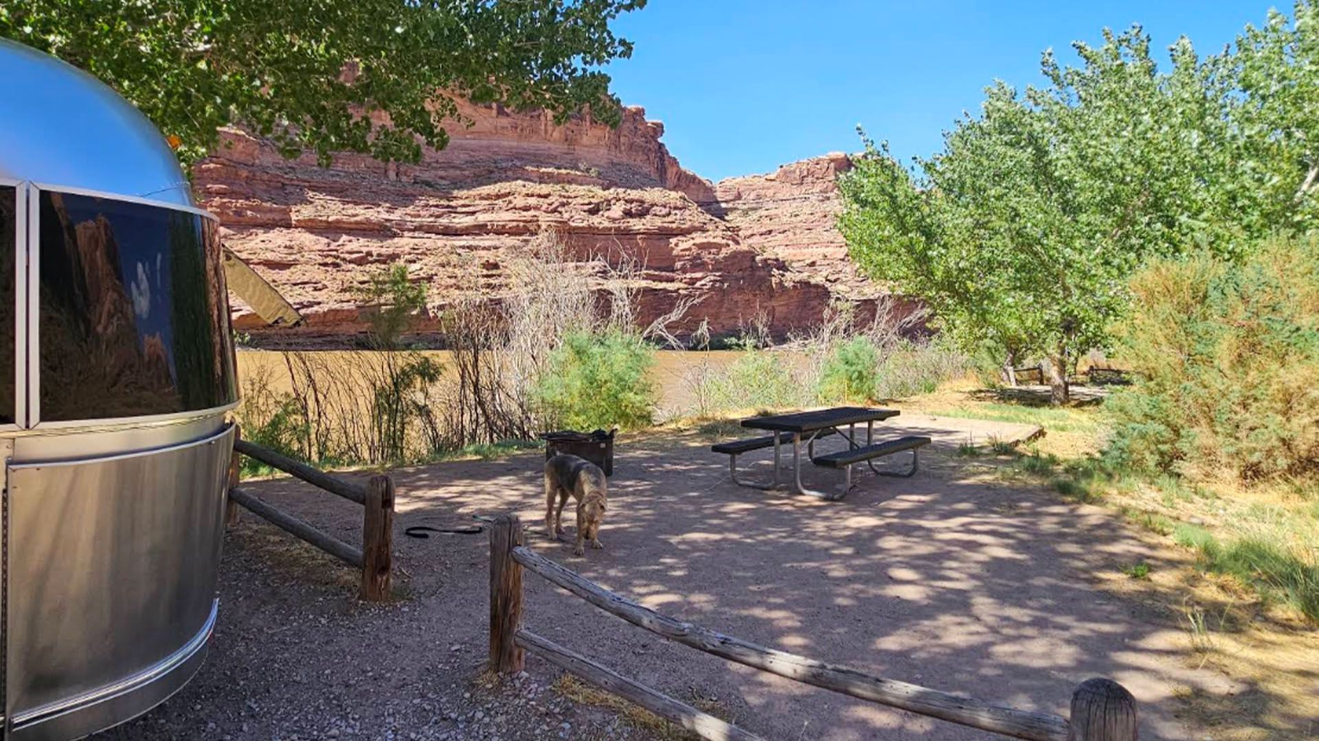 Silver camper parked at a campsite, with a picnic table and red rock cliffs in the background on a sunny day.