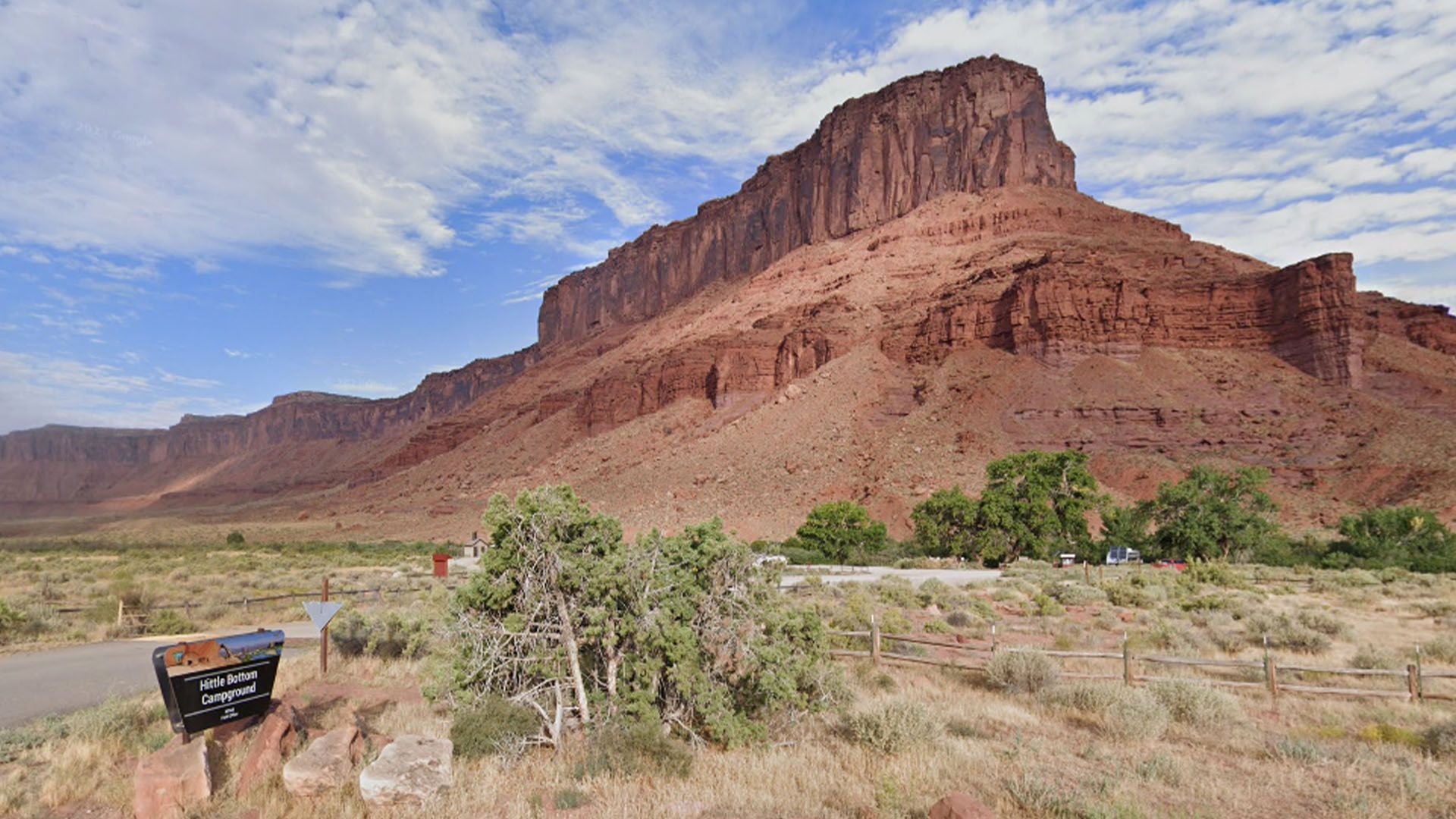 Red rock mesa in desert landscape with a blue sky and low bushes next to a campground entrance sign.