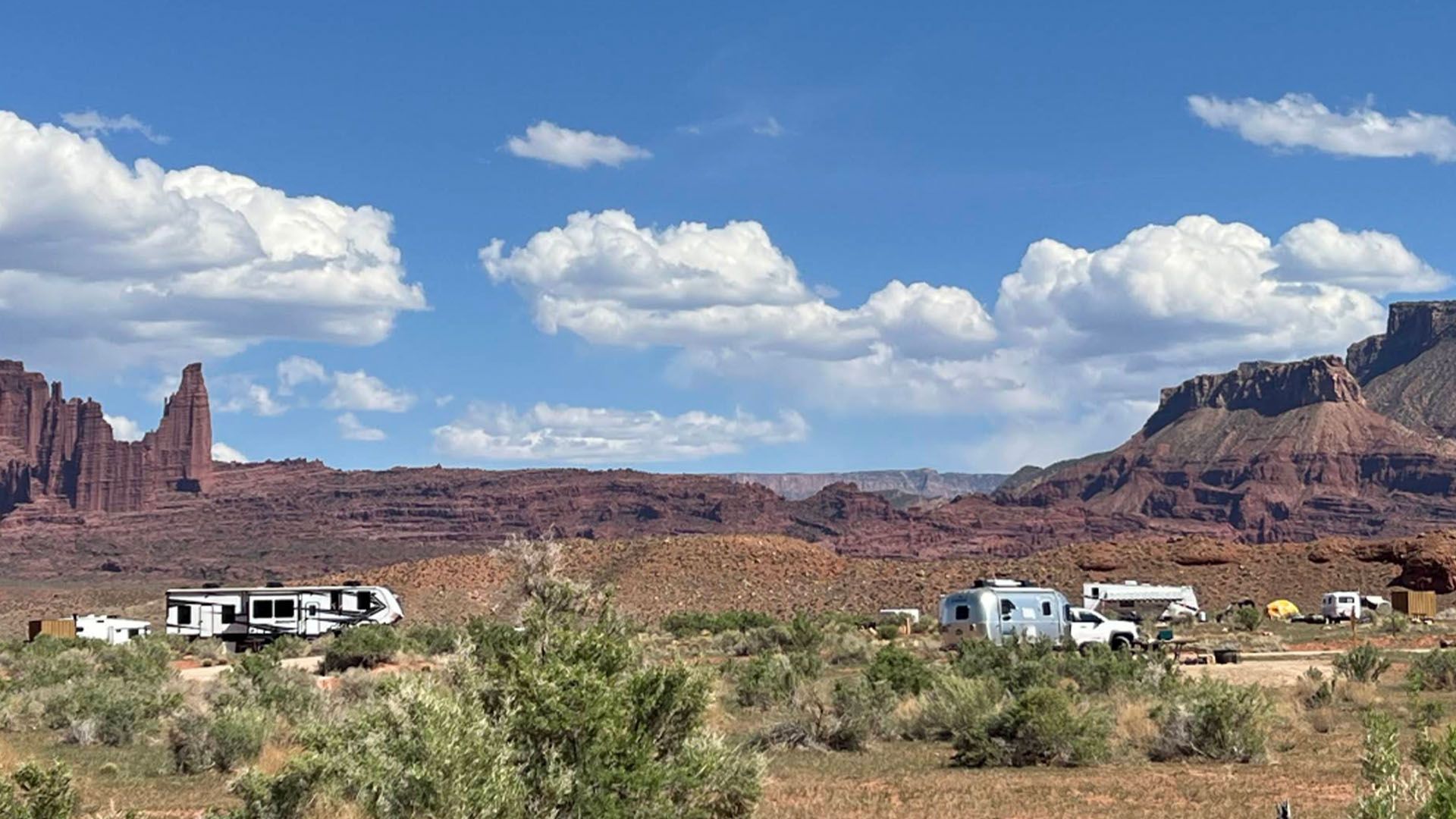 Camping area with RVs amidst red rock formations under a blue sky with puffy clouds.