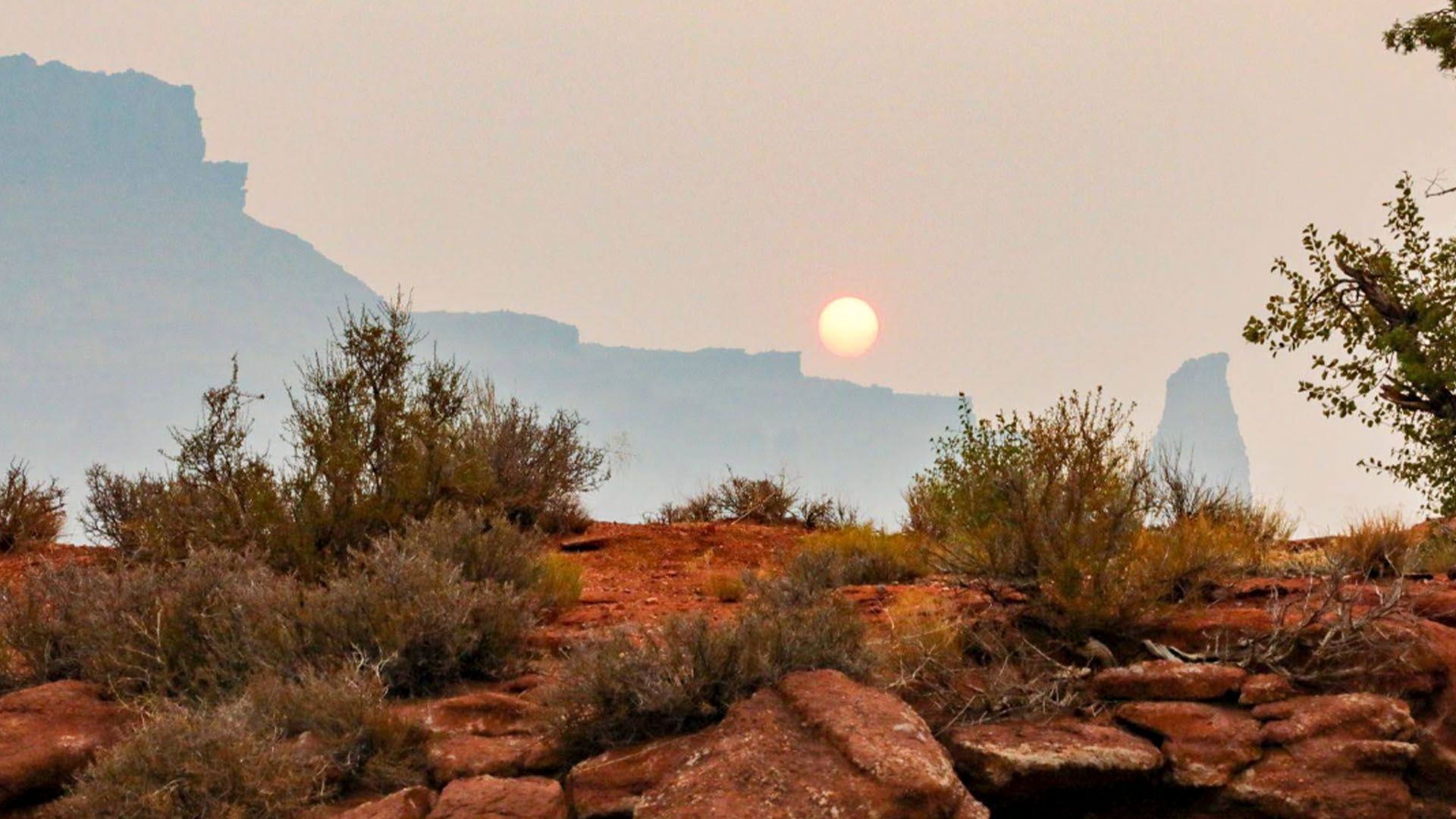 Sun obscured by haze over a desert landscape with red rocks and scrub brush.