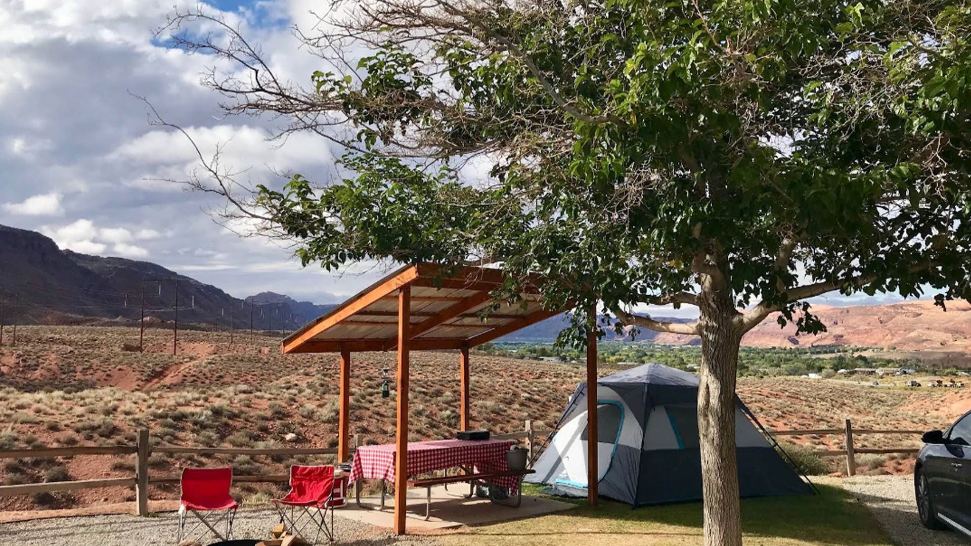 Camping scene with tent, picnic table under shelter, red chairs, desert landscape.
