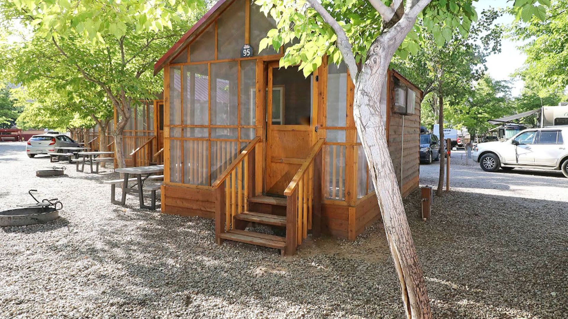 Wooden cabin with screened porch, steps, trees, and gravel ground.
