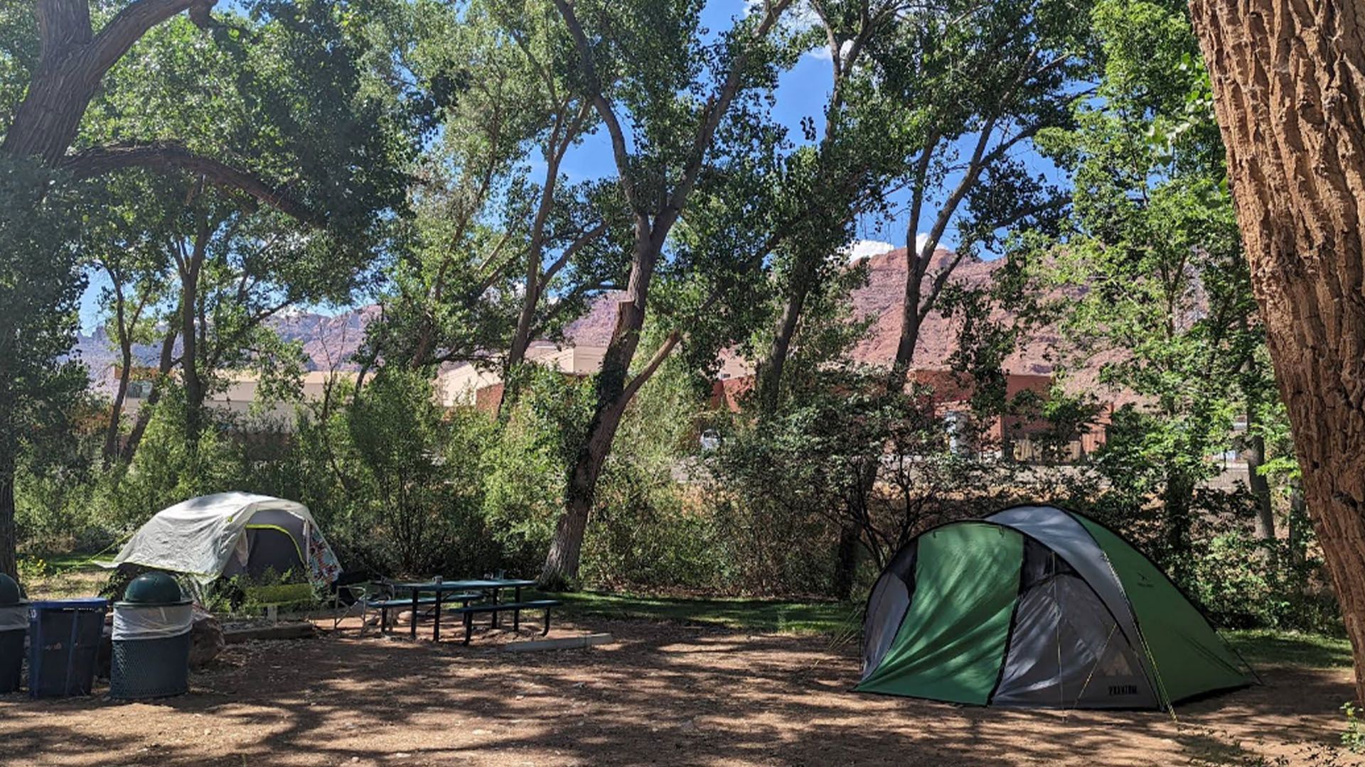 Campsite with two tents, a picnic table, and trash cans under trees. Red cliffs are visible in the background.