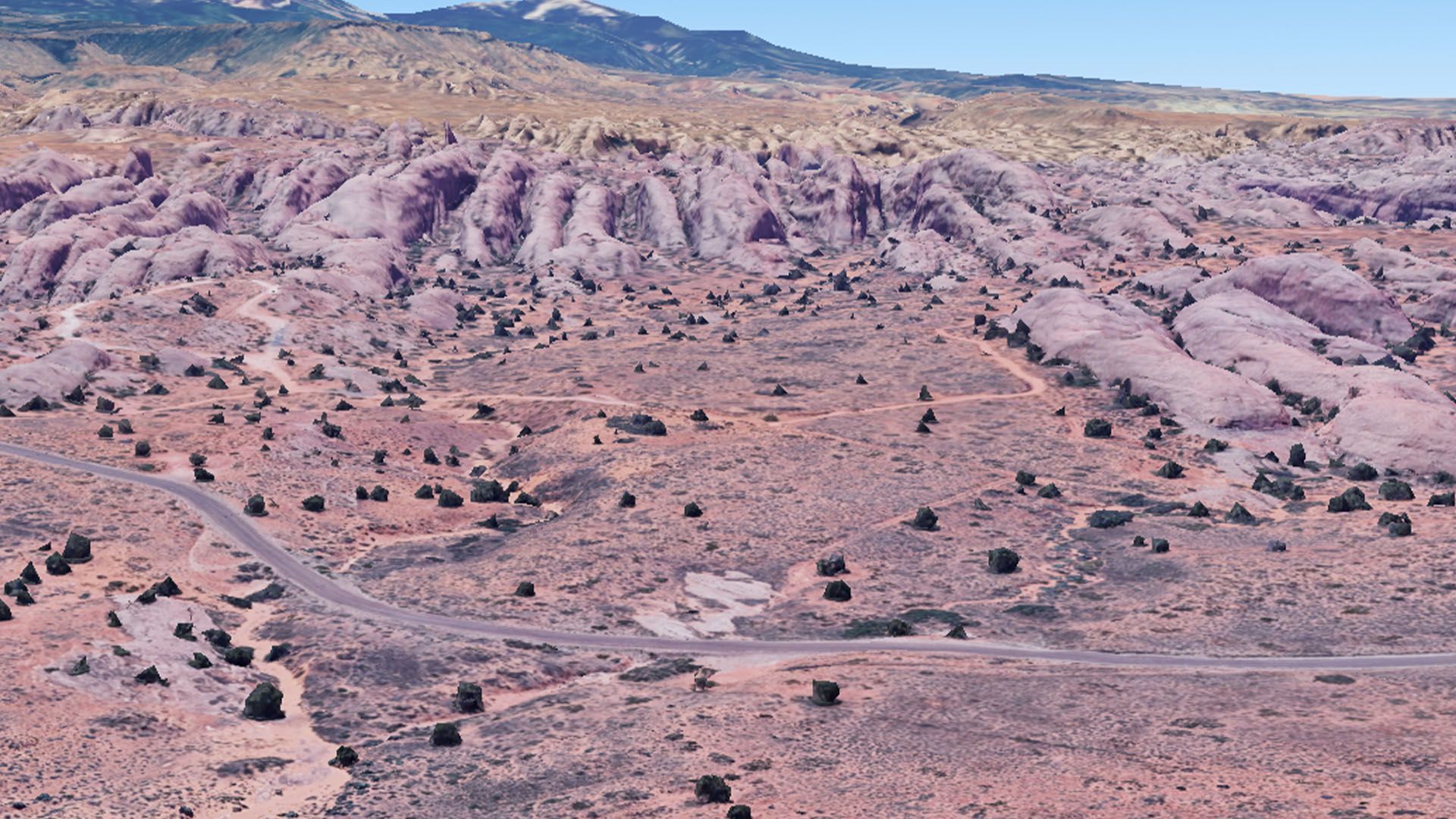 Desert landscape with reddish-brown rock formations, sparse vegetation, road winding through, and distant mountains.