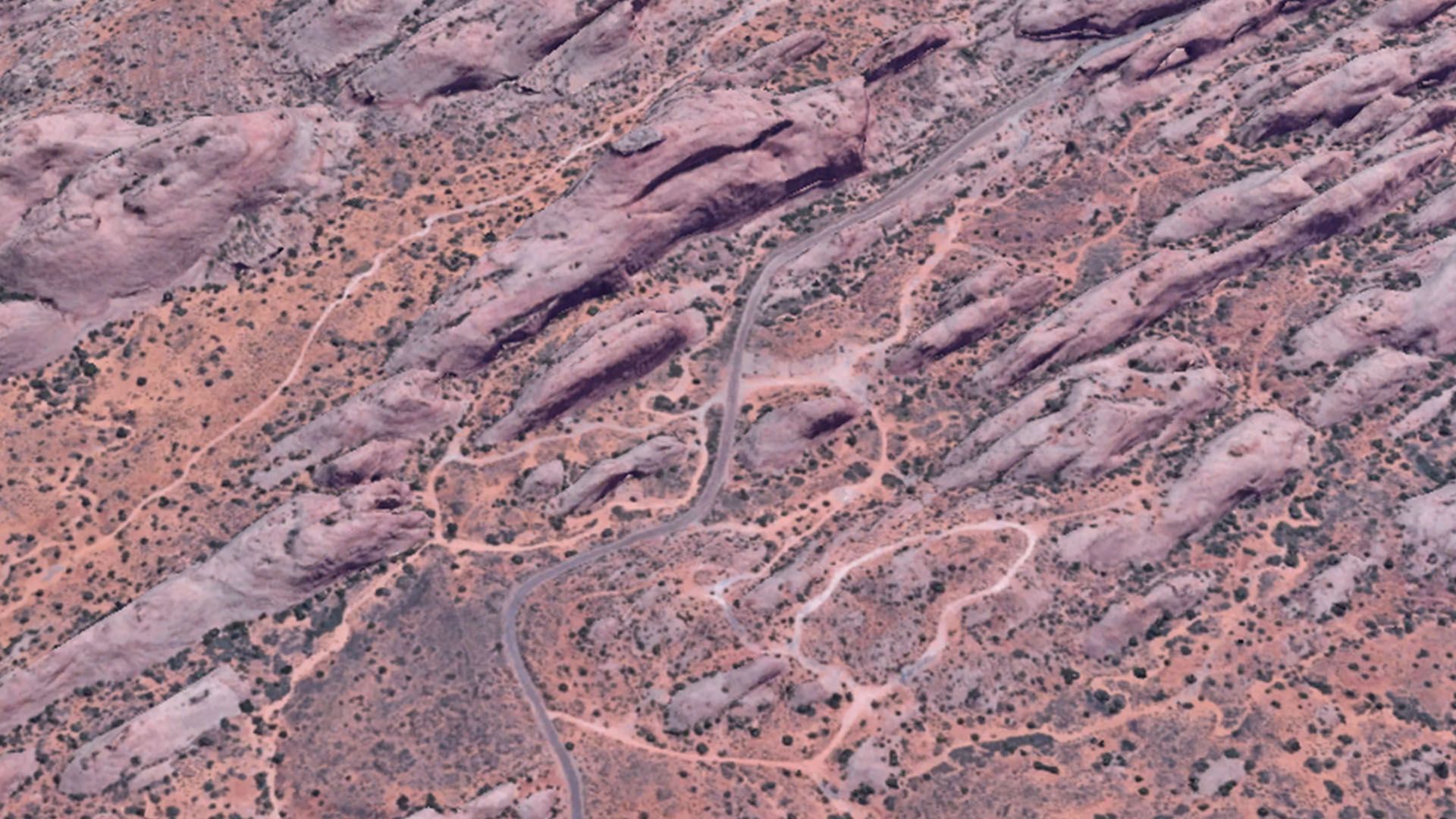 Aerial view of a desert landscape with a winding road and trails through red rock formations and sparse vegetation.