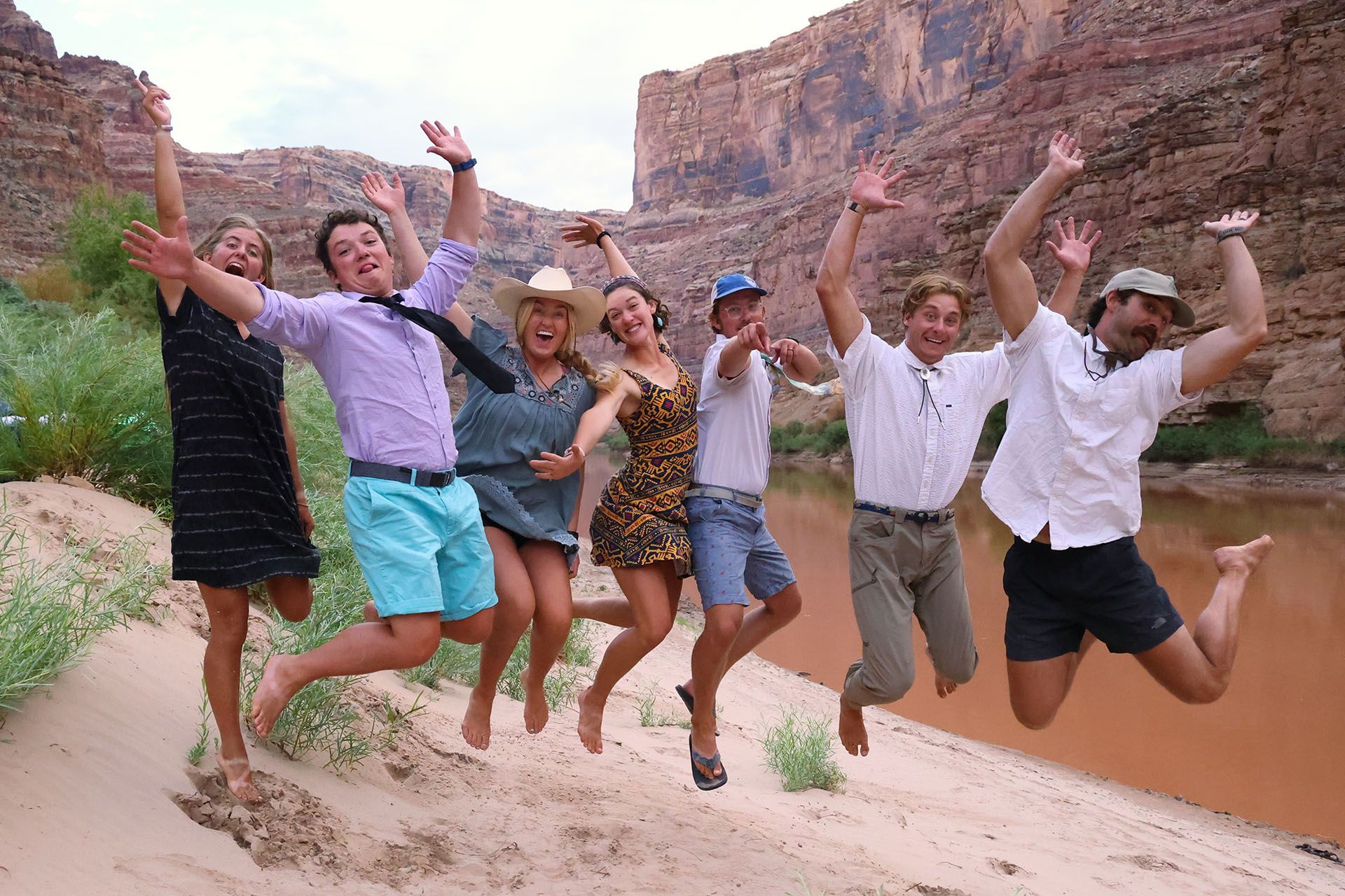 Group of people jumping joyfully on sandy bank by a red river, with canyon backdrop.