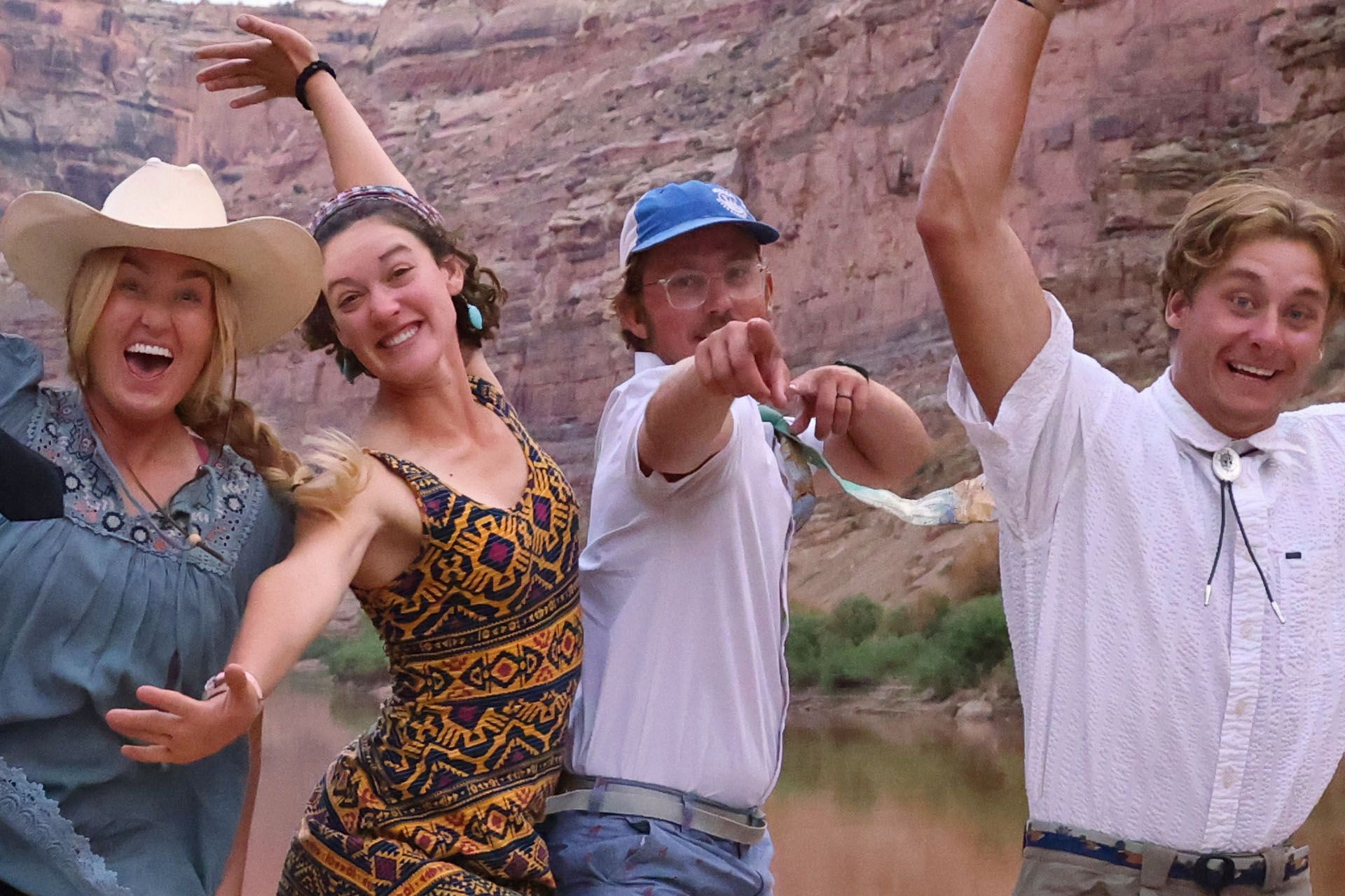Four people pose joyfully near a river, canyon backdrop.