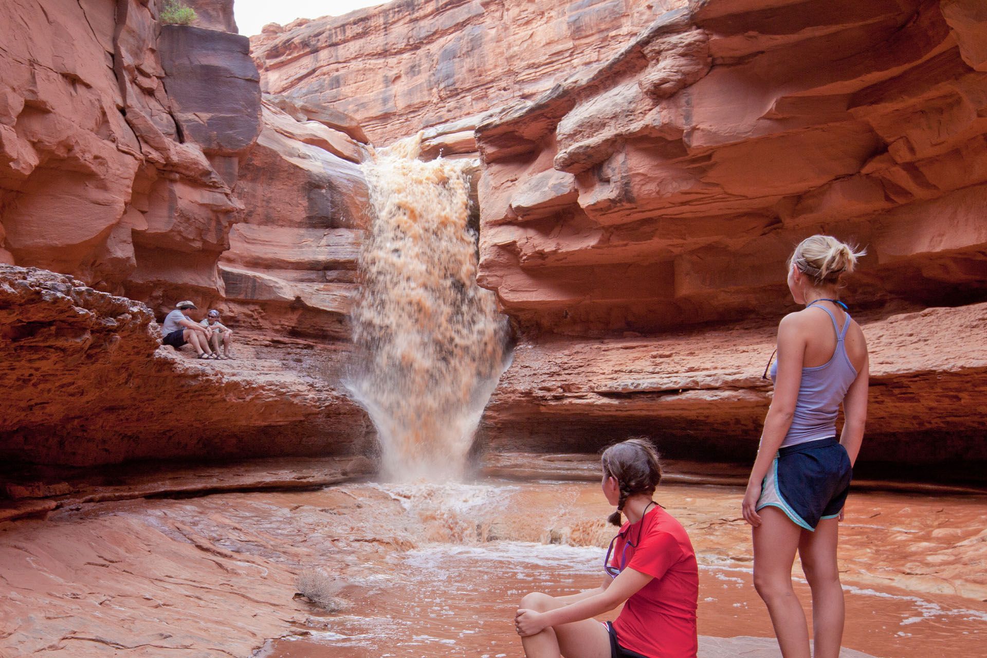 Two girls look at waterfall pouring over sandstone cliff.