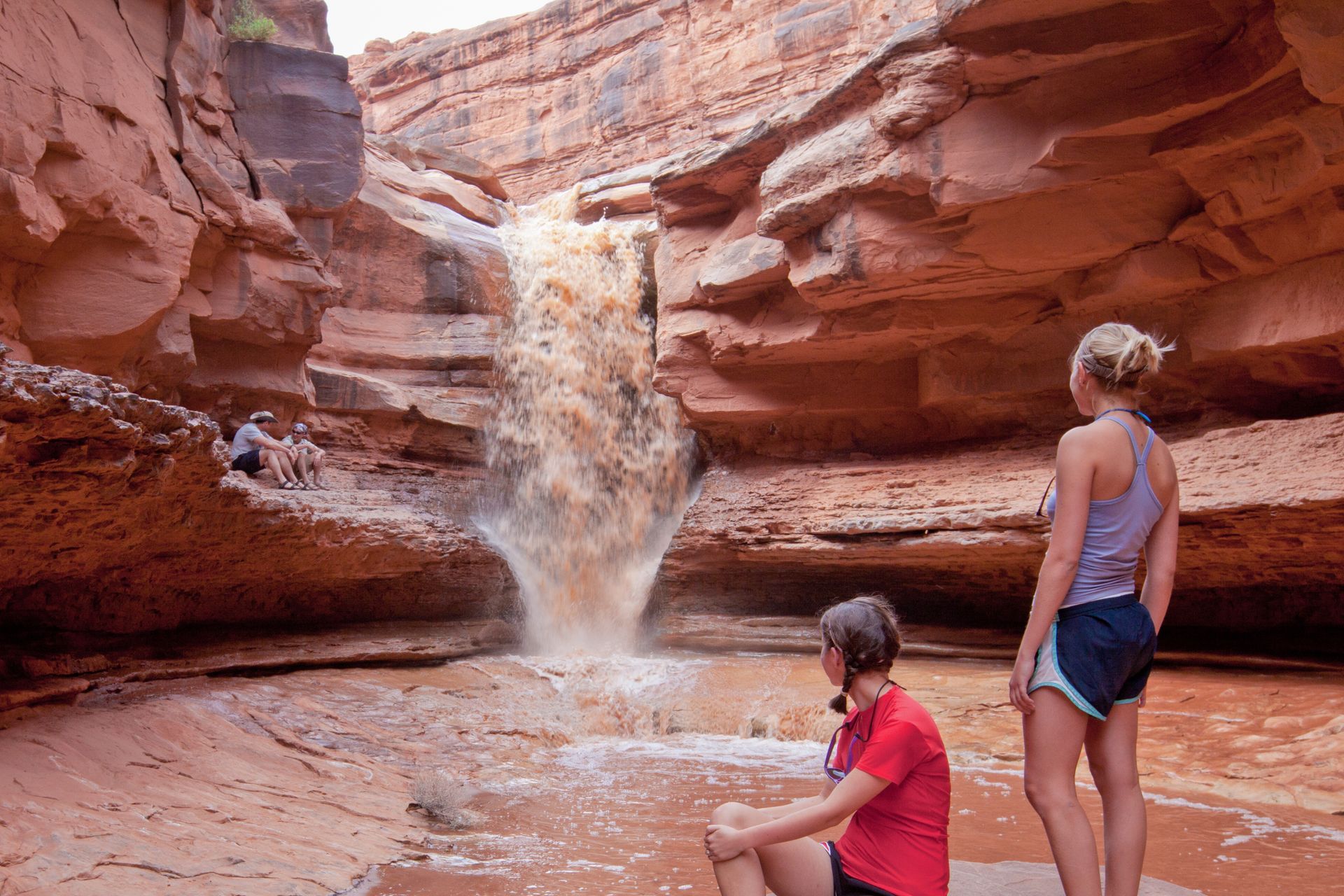 Two people standing in front of Indian Creek Falls with water pouring off the top of the cliffs and down the canyon.