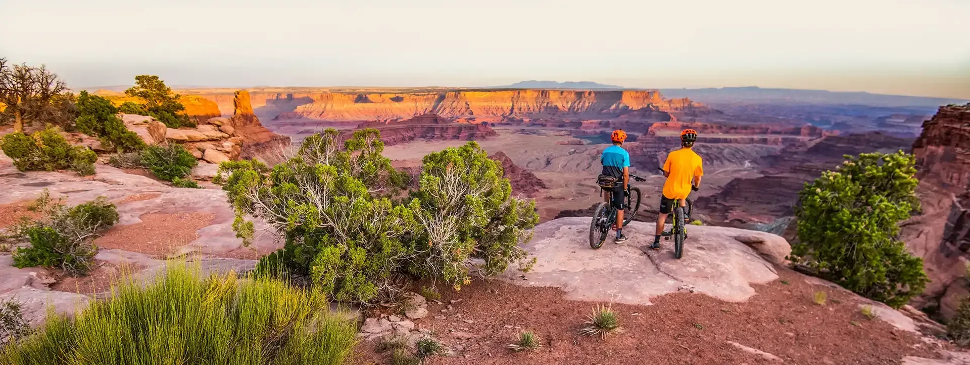 Two cyclists overlooking a vast canyon at sunset. They are standing on a rocky outcrop next to their bikes.